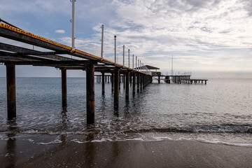 Fototapeta premium Vintage Beach Pier Extending into the Sea. Old Metal Pier on a Beach Resort Coastline. Abandoned Iron Pier Along the Shoreline.