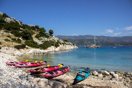 Vibrant horizontal shot of red and cyan sea kayaks parked on a white rocky shore in Kekova bay, Turkey. Clear turquoise water and a cruising yacht visible in the background under a blue sky.