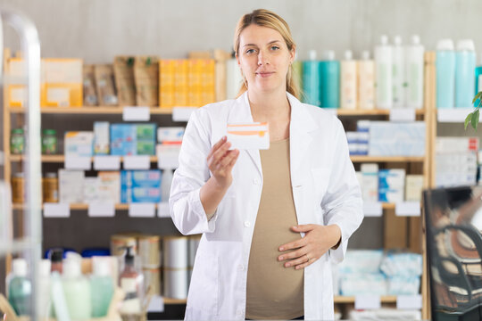 Friendly pregnant apothecary holding a box with remedy standing at the desk in drugstore