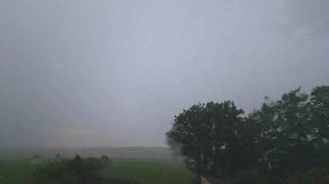 View through window showing heavy falling rain streaks during an intense thunderstorm with strong winds affecting trees over a green rural landscape near Looe, United Kingdom.