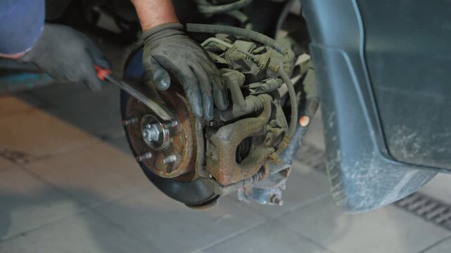Close up view of a car mechanic's hands in gloves cleaning a rusty brake disc hub with a wire brush during vehicle maintenance in an auto repair shop, preparing for brake pad replacement