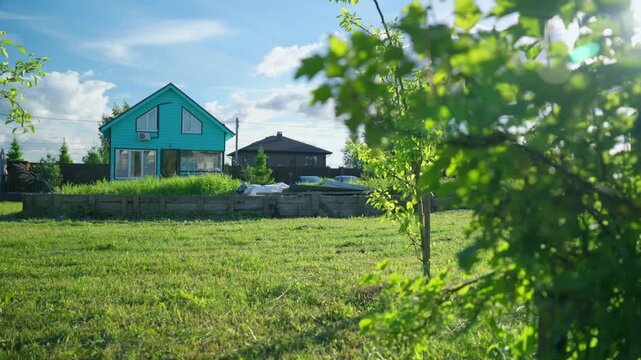 Bright turquoise cottage standing in a rural area with a lush green lawn and garden beds, viewed from behind swaying tree leaves on a beautiful summer day with a clear blue sky and clouds