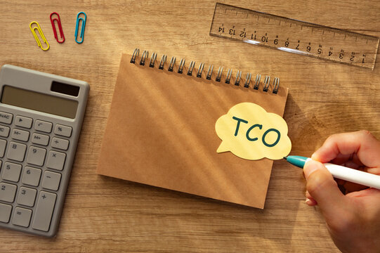 TCO total cost of ownership &mdash; financial business concept with hand writing on sticky note, bright wood desk with calculator