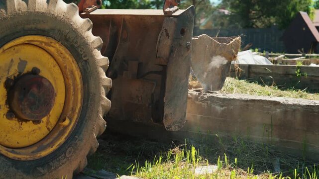 Close-up of backhoe loader wheel and bucket beside an old concrete foundation in a residential yard, showing demolition equipment, site preparation and groundwork.