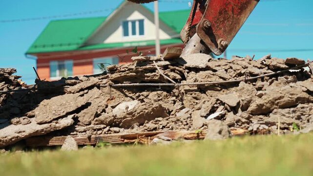 Industrial hydraulic hammer breaking reinforced concrete and asphalt into rubble at a demolition site. Heavy machinery working on a construction project against a suburban house background