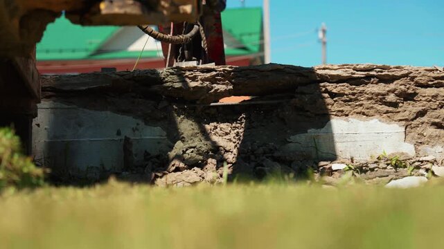 Heavy machinery with a hydraulic hammer demolishing a concrete and stone wall, creating debris and dust during the destruction process on a bright and sunny day at a construction site