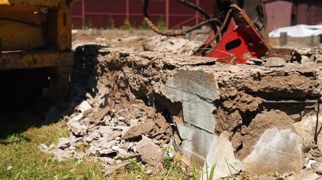 Powerful excavator with a hydraulic crusher attachment breaking apart and demolishing a reinforced concrete foundation, creating a pile of rubble and debris on a construction site
