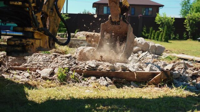 Backhoe loader bucket crushing concrete rubble and dust in a residential yard, showing demolition cleanup, foundation removal and site preparation for new construction.