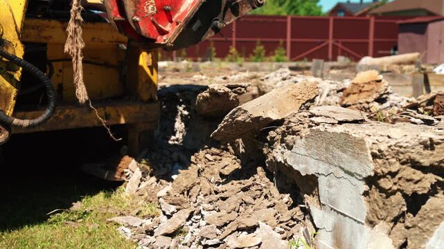 Industrial demolition machine with a rotating milling head breaking down an old concrete foundation, creating debris and dust at a construction site during a sunny day
