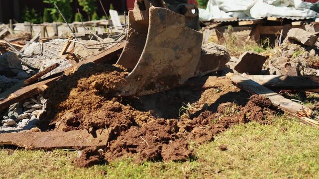 Close-up of backhoe loader bucket spreading soil over demolition debris and broken foundation rubble in a residential yard, showing site cleanup and groundwork.