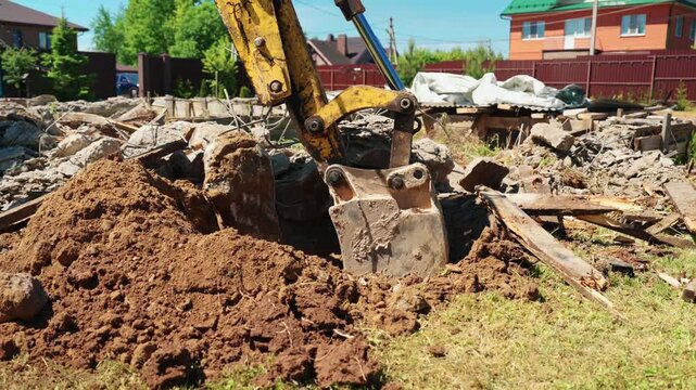 Close-up of backhoe loader bucket digging soil among concrete rubble and broken wooden debris in a residential yard, showing site cleanup and groundwork.