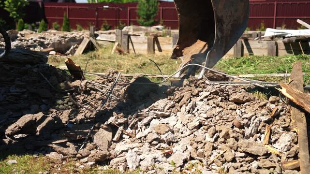 Close-up of backhoe loader bucket sifting concrete rubble and exposed rebar from demolished foundation debris, showing site cleanup and residential preparation.