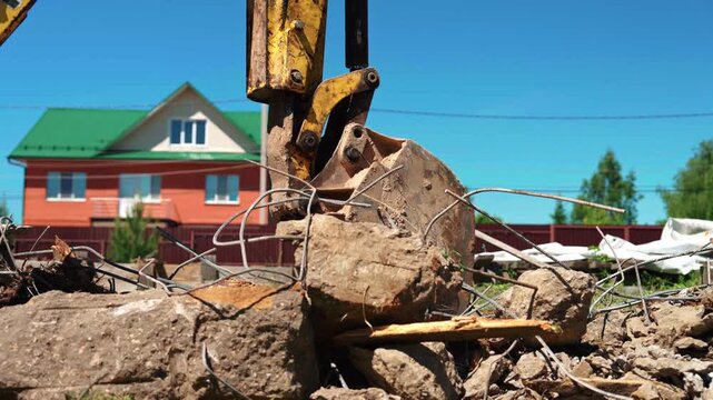 Heavy machinery at a construction site demolishing reinforced concrete blocks, with twisted rebar sticking out from the rubble under a clear blue sky, while a house stands in the background