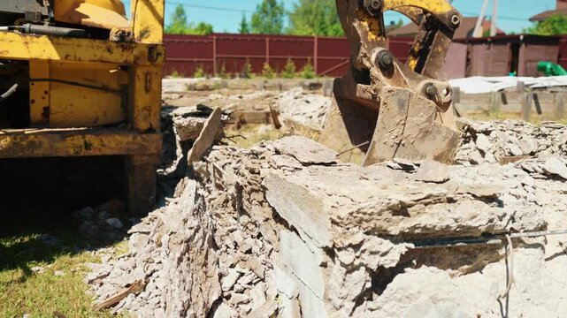 Close-up of backhoe loader bucket clearing rubble from a broken concrete foundation with exposed rebar, showing demolition cleanup and residential site preparation.