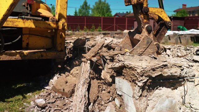 Powerful yellow excavator with a bucket attachment breaking up reinforced concrete foundation at a construction site during demolition work, with debris and rubble flying around on a sunny day