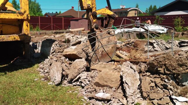 Heavy machinery dismantling a reinforced concrete structure at a building site during demolition work. The excavator's bucket crushes stone, rubble, and metal rebar with powerful force