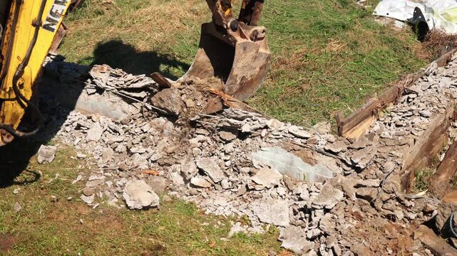 Backhoe loader bucket dropping soil onto concrete rubble and broken wood from an old foundation, showing demolition cleanup, debris handling and site preparation.