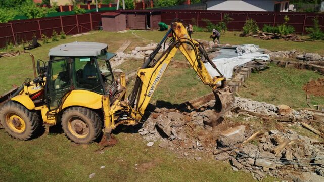 Powerful yellow backhoe loader breaking up and removing an old concrete foundation with its bucket during demolition work on a residential construction site, seen from an aerial view