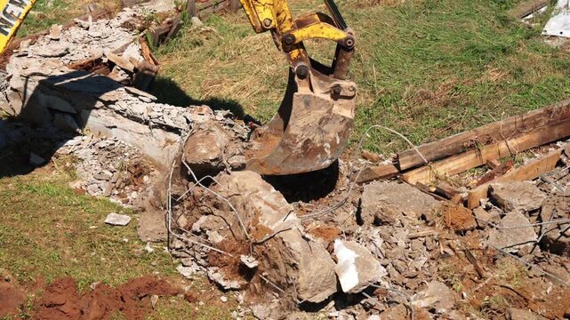 Backhoe loader bucket clearing concrete rubble, broken wood and exposed wires from an old foundation trench, showing demolition cleanup and residential site preparation.