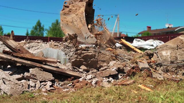 Close-up of backhoe loader bucket scraping exposed rebar, concrete rubble and broken wood from a demolished foundation, showing site cleanup and residential preparation.