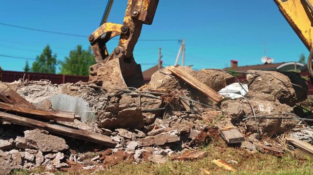 Powerful yellow excavator arm with a bucket actively demolishing an old concrete foundation, breaking it into rubble and debris on a construction site under a clear blue sky during the daytime