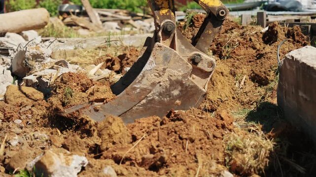 Close-up of backhoe loader bucket scooping soil from demolished foundation debris with concrete rubble, broken wood and exposed rebar during residential site cleanup.