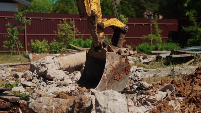 Close-up of backhoe loader bucket sorting concrete rubble, exposed rebar and broken wood in a residential yard, showing demolition cleanup and site preparation.