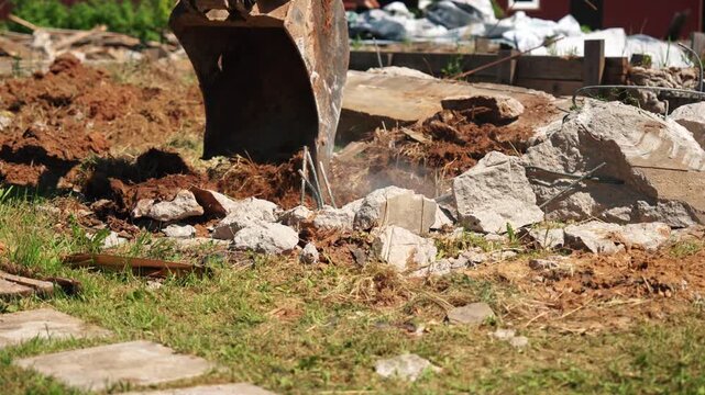 Heavy machinery with a powerful digging bucket breaking apart an old, reinforced concrete slab during demolition work at a residential construction site, raising dust and debris