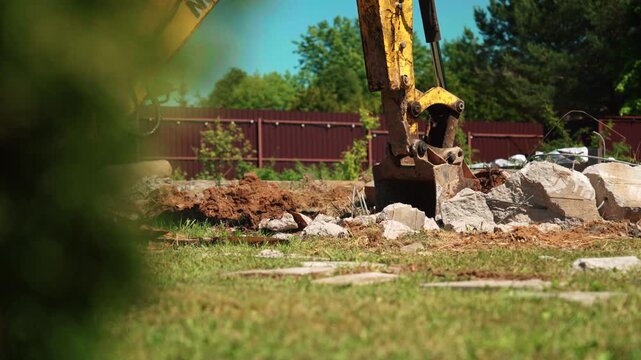 Heavy machinery excavator with a hydraulic bucket actively demolishing a concrete foundation during construction work. Industrial equipment breaking large stones and debris in a residential backyard