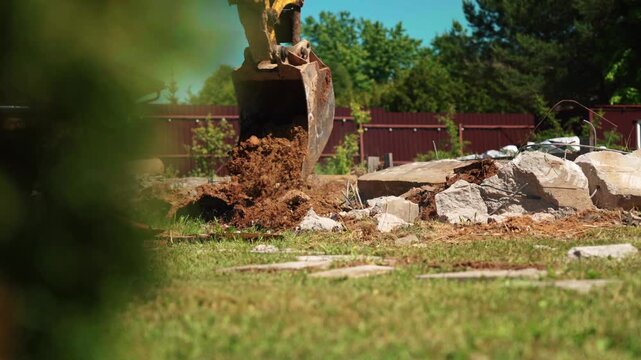 Backhoe loader bucket dumping soil beside broken concrete rubble and exposed rebar in a residential yard, showing demolition cleanup and site preparation.