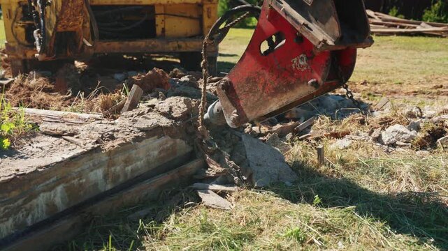 Close-up of a hydraulic breaker resting on a reinforced concrete foundation beam with exposed rebar before demolition, showing heavy equipment, structural removal and residential site preparation.