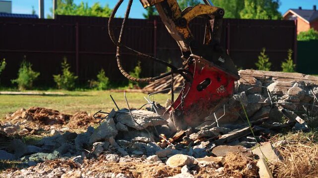 Powerful hydraulic breaker attached to an excavator smashing reinforced concrete into small pieces on a sunny day. Construction machinery is demolishing a structure, creating dust and rubble