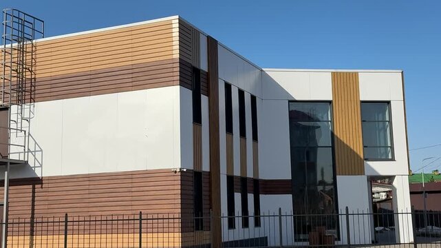 Stylish modern house with siding under blue sky, geometric facade of glass and wood panels, parking lot and metal fence