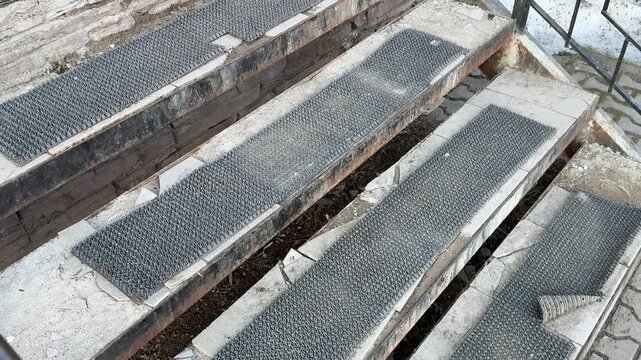Worn antiskid coating on steps at pickup entrance, rusted metal treads with peeling paint and grime, uneven concrete pavers, damp