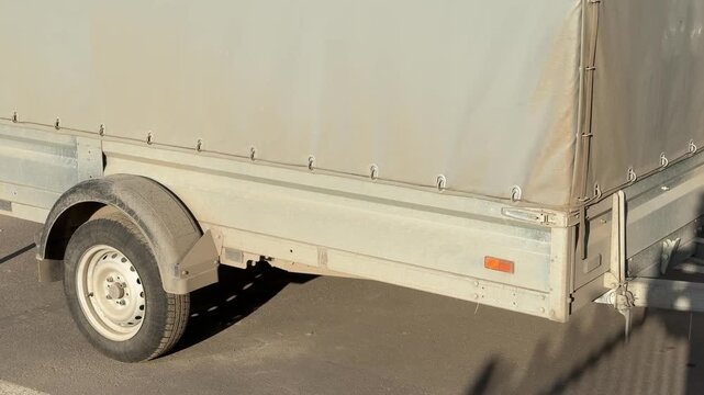 Covered passenger trailer parked in lot during spring sunlight gray tarpaulin fastened over cargo bay single axle wheel, hitch