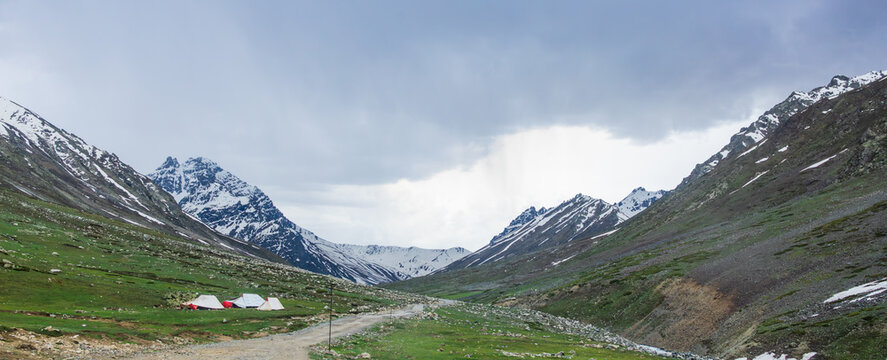 A rugged dirt road leads toward a small base camp nestled within a dramatic snow capped mountain range, remote expedition camp pitched in a vast Himalayan valley, 