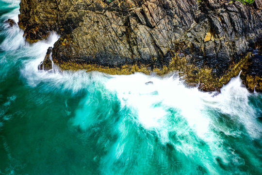 Aerial view of waves crashing against the rugged dark rocks of the coastline with turquoise water and white sea foam at Cabarita Beach, New South Wales, Australia.