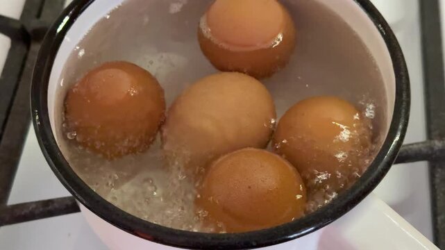 Close-up boiling eggs in iron ladle on gas stove, five brown shells arranged in water, gentle simmer with small bubbles, enamel