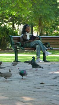 Vertical video: Approaching park bench, woman sitting and reading book to study with pigeons nearby