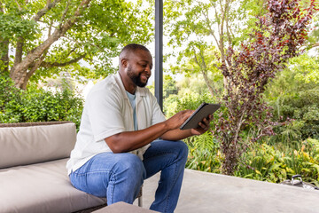 Fototapeta premium African American man sitting on covered patio sofa using tablet, wearing shirt and jeans near post