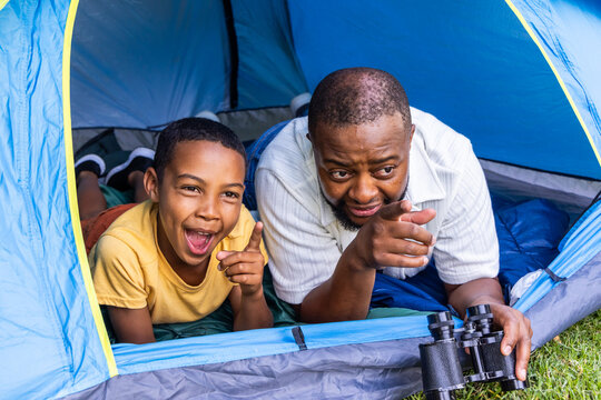 African American father and teen son lying on stomachs at blue tent, holding binoculars, pointing