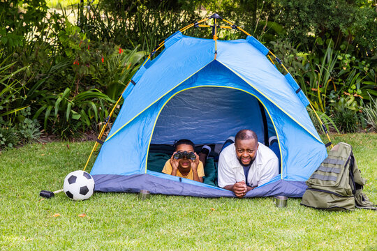 African American male father and child lying in backyard tent using binoculars beside backpack