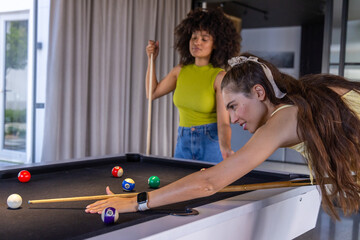 Fototapeta premium Diverse female friends leaning in game room black-felt pool table, aiming cue near striped balls