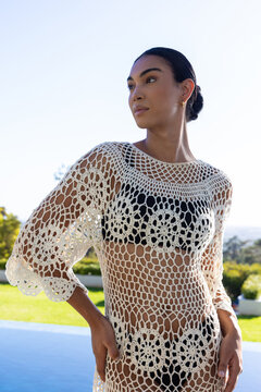 Adult African woman posing poolside wearing white crochet coverup over black swimsuit, gazing sea