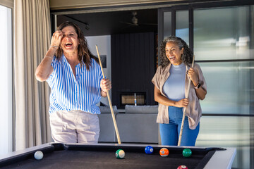 Naklejka premium Diverse female friends laughing, playing pool with cues, billiard balls near sliding glass doors