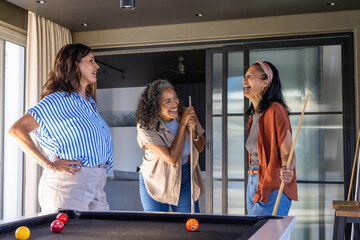 Naklejka premium Diverse female friends laughing, playing at pool table in home rec room, holding cue sticks