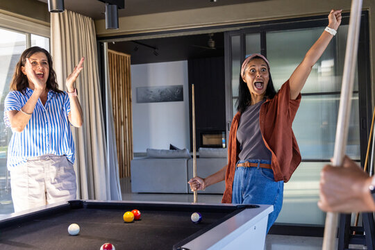 Diverse mature female friends cheering and clapping at home by pool table with billiard balls