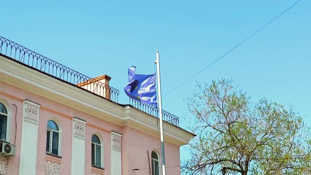 European Union flag waving on a flagpole against a blue sky. The blue flag with yellow stars of the European Union flutters in the wind on top of a classic pink building facade. 