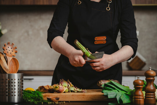 Woman private chef peeling thick stem of raw artichoke with knife. Professional vegetable cleaning technique and organic culinary arts concept.