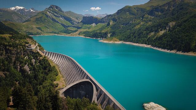 Aerial view of the Roselend Dam with its curved concrete structure holding back the turquoise waters of Lac de Roselend, surrounded by mountains in Beaufort, Auvergne-Rhone-Alpes, France.